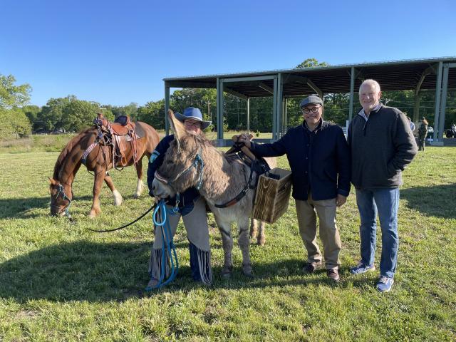A donkey wearing a wooden pack saddle stands with an adult handler, while two other adults stand beside them; behind them, a saddled horse grazes and a large open-sided structure sits in the background.