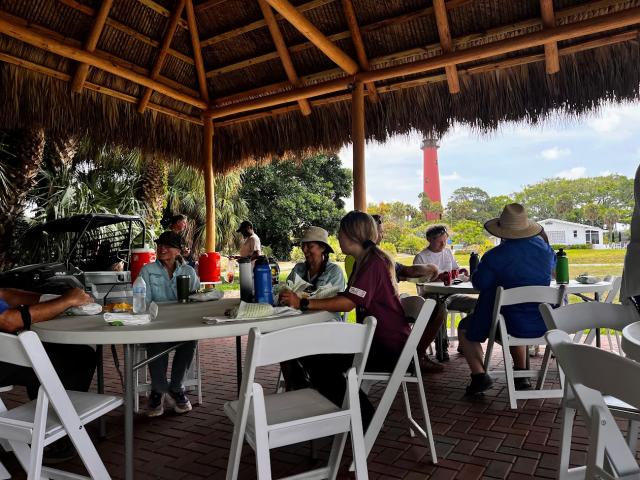 Group of people sitting and chatting under a thatched roof pavilion with a red lighthouse visible in the background.