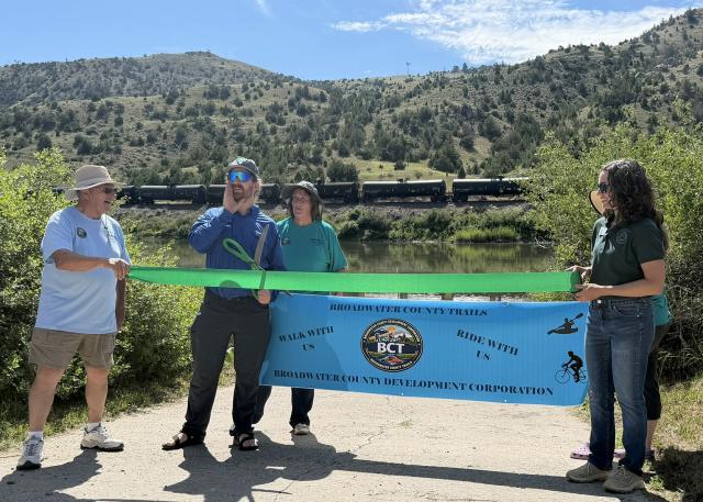People hold a green ribbon on a boat launch with hills in the background as a person cute it with big scissors