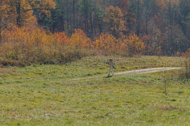 Person dressed in colonial attire walking along a path in a grassy field with autumn-colored trees in the background.