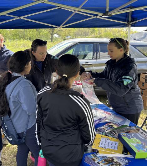 A volunteer in a uniform hands out supplies to three women under a blue canopy at an outdoor event.