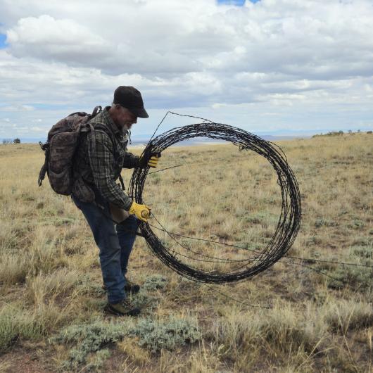A man has a large old barbed wire fence in his hand in a large circle with clouds in the sky and brown grass. 
