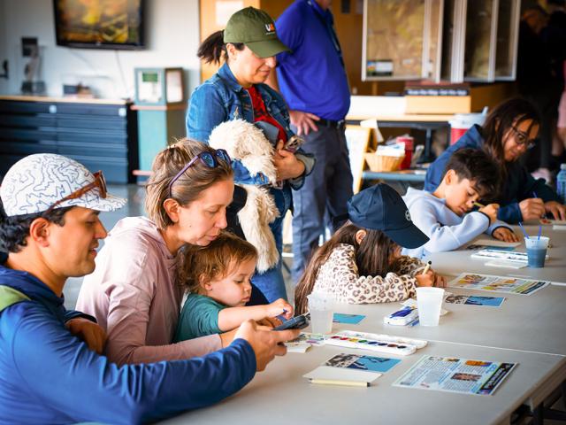 A group of adults and children sit at long tables indoors, participating in an arts-and-crafts activity. Several children are focused on coloring or painting, while adults nearby assist or observe. In the background, a person stands holding a fluffy white jacket, and others move around the room. Various supplies (cups of water, paint palettes, papers) are spread across the tables, creating a busy, creative atmosphere.
