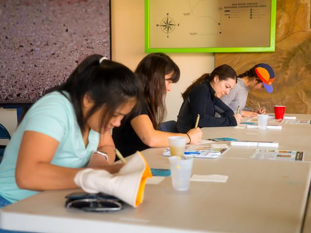 Several people sit at a long table indoors, each focused on creating artwork. They lean over postcard-sized pieces while drawing or painting, with cups of water, art supplies, and reference sheets scattered across the table. A large wall map and display behind them indicate the activity is taking place in a visitor or education center.