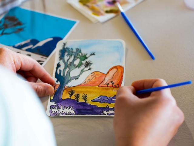 A person holds a small postcard-sized watercolor painting depicting a desert landscape with orange rock formations, a large tree in the foreground, and purple and yellow plants along the ground. The person uses a blue paintbrush to add details, with other painted cards and supplies visible on the table.