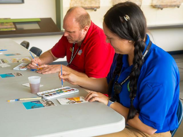 Two people sit at a table indoors, each painting small postcard-sized artworks with watercolor palettes. One person in a red shirt concentrates on adding color to a postcard, while the person beside them in a blue shirt smiles as they paint. Various art supplies, water cups, and partially completed postcards are spread across the table.