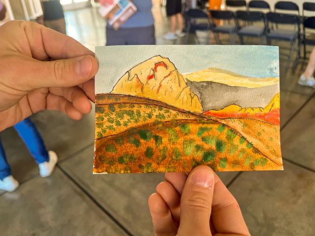 A person holds a small watercolor postcard with both hands. The painting shows a landscape of golden and orange rocky hills, green vegetation in the foreground, and a soft blue sky above. The background of the photo is slightly blurred, showing people standing and folding chairs in an indoor setting.