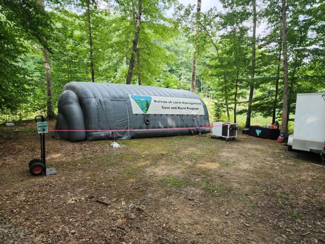 A large gray inflatable tunnel structure sits in a wooded outdoor area surrounded by tall green trees. A banner on the tunnel reads “Bureau of Land Management – Cave and Karst Program.” A red rope line marks the entrance area, with a small sign indicating where to enter. Nearby are equipment cases, a table with supplies, and a white trailer. The ground is covered in dirt and leaves.