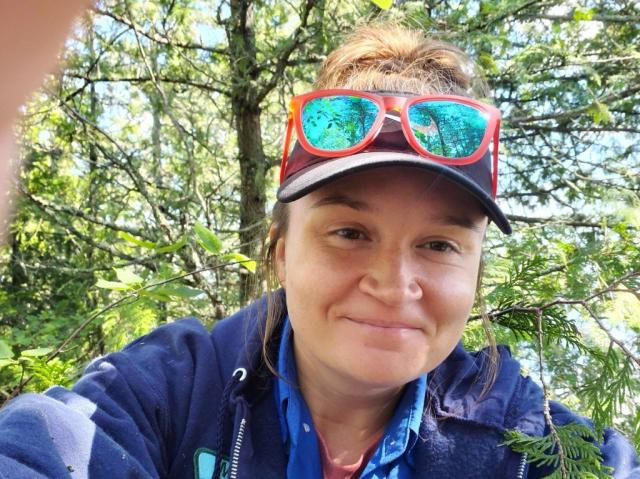 A person outdoors wearing a cap and bright red sunglasses perched on top of the hat, smiling slightly while surrounded by sunlit trees and branches.