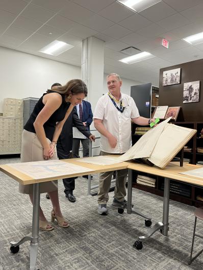 Two people stand at a table examining a large open book of historical maps or documents. One person leans in closely to look at the pages while another gestures toward the material. Additional observers and filing cabinets are visible in the background.