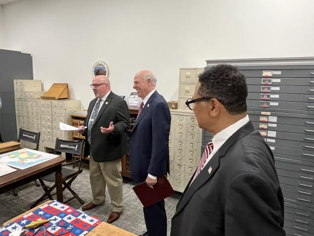 Three people stand together in a room with map drawers and archival furniture. One person speaks while holding papers, and the others listen attentively. Colorful documents and historical materials are laid out on nearby tables.