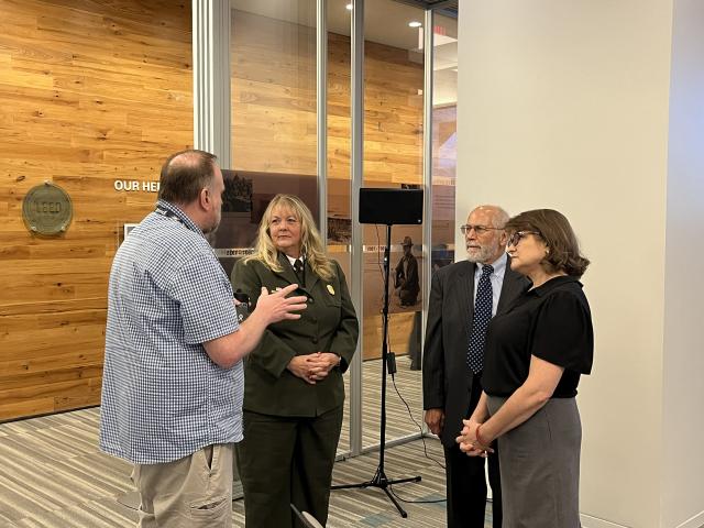 Four people stand in conversation inside a modern building with wood-paneled walls and historic interpretive displays behind glass. One person gestures while speaking, and the others listen.