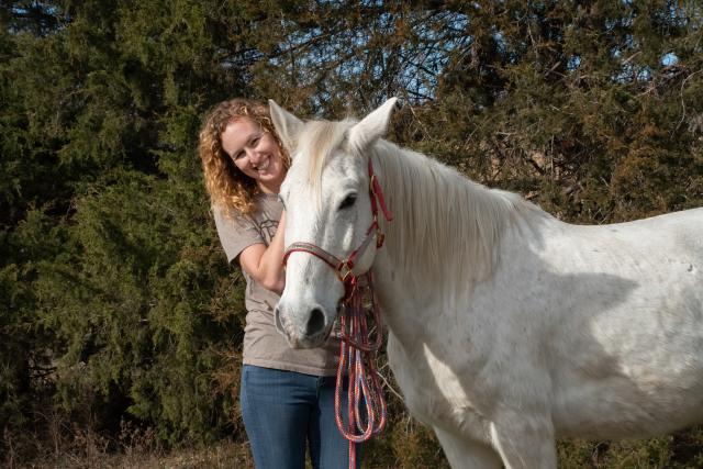 A photo showing a white horse looking into the camera with Lindsey Gabbard standing next to him, smiling.