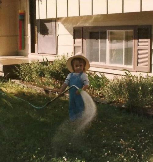 A small child standing in a yard, wearing overalls and a wide-brimmed hat, holding a garden hose that sprays water across the grass near a house.