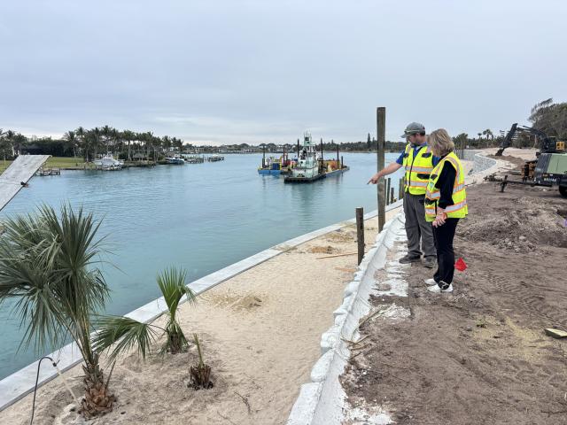 Two people wearing high‑visibility vests stand on a newly constructed shoreline edge, looking over calm water where barges and equipment are present.