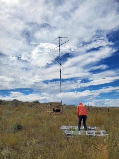 A person in an orange hooded jacket stands beside metal tower sections laid on the ground in a grassy, sagebrush-covered area. A tall antenna mast with multiple horizontal elements rises overhead against a blue sky filled with layered white clouds.