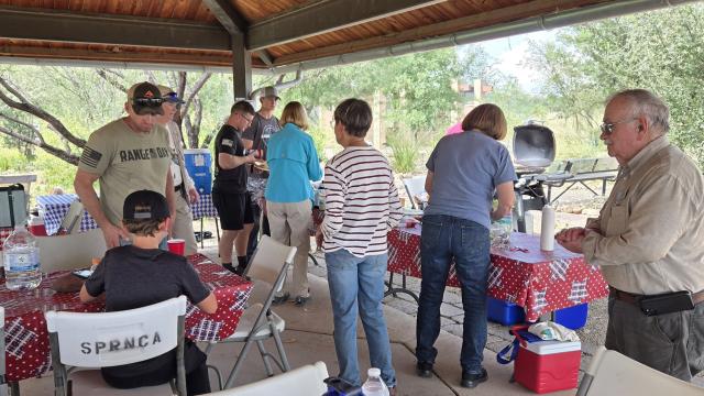a group of people under a shade structure with picnic tables and grills