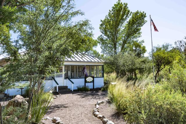 a ranch house with a flag flying outside. The house is surrounded by green desert vegetation and tall trees.