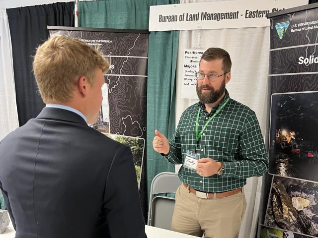 Two men converse at a Bureau of Land Management booth displaying posters and information about careers and mining.