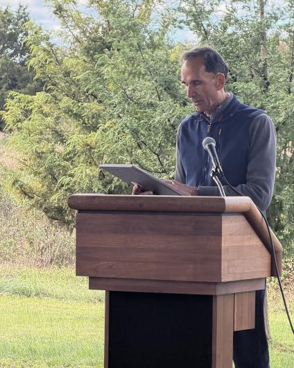 A speaker stands at a wooden podium outdoors, reading from a plaque into a microphone with trees and natural vegetation in the background.