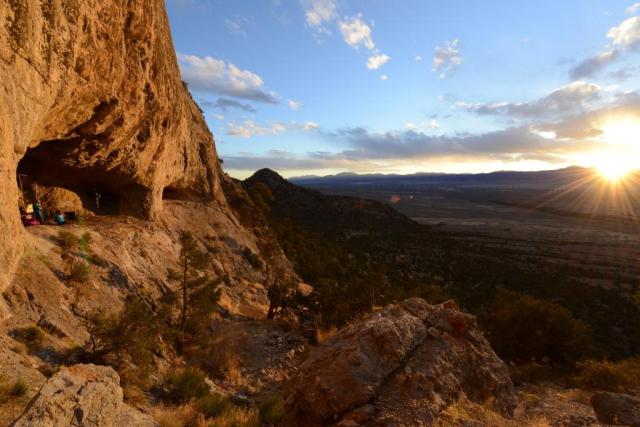 A rocky cave entrance opens along a steep cliff face overlooking a wide desert valley at sunset. Golden sunlight illuminates the rugged rock wall and foreground boulders, while distant mountains form a dark silhouette against the horizon. The sun is low in the sky, casting long shadows across the landscape and highlighting the dramatic terrain surrounding the cave.
