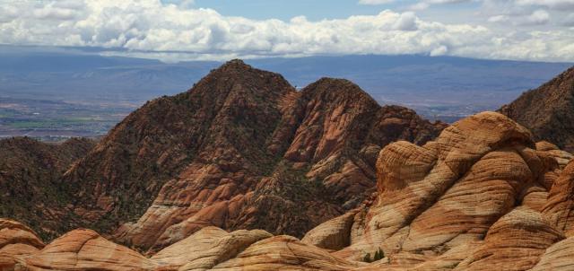 Layered red and beige formations in the foreground with rugged mountain peaks under a cloudy sky in the background.