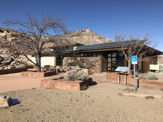 A small visitor center-style building with stone and stucco walls sits at the base of rocky desert hills. The building has a low roof with solar panels, and leafless trees and desert shrubs surround the paved walkway. An accessibility sign and informational display panels stand near the entrance.