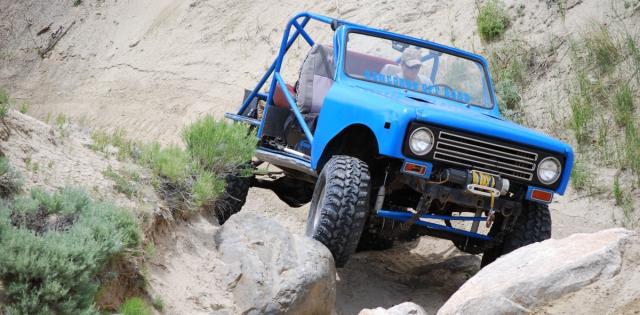 A bright blue off‑road vehicle with large tires navigates over rocks and a steep sandy slope in a rugged, desert-like landscape with sparse shrubs.