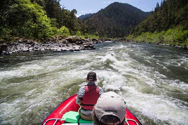 The view from behind two people in a red raft heading down a river toward whitewater rapids.The view from behind two people in a red raft heading down a river toward whitewater rapids. Both wear caps, and the person in front wears a life vest. The river is flanked by steep, forested mountains and rocky banks under a blue sky.