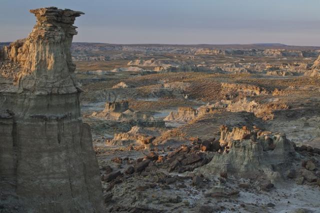A wilderness study area landscape with eroded rock formations at golden hour. A tall hoodoo with a flat, overhanging cap rises in the foreground on the left, while smaller rock spires and weathered formations dot the expansive valley beyond. The scene is bathed in warm light under a pale sky.