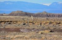 A solitary pump jack or oil well structure stands in a wide, open desert landscape. The terrain is flat and arid with sparse vegetation, scattered rock formations, and low hills or mesas in the distance under a pale sky.