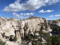 Eroded rock formations and cone-shaped hoodoos rise from a desert landscape under a partly cloudy blue sky, with sparse trees in the foreground.