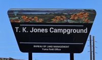 A trapezoid-shaped sign displays a landscape banner with flowers in the foreground and a lake in the background. Centered text below the banner reads “T.K. Jones Campground.” Along the bottom of the sign are two lines of text: “Bureau of Land Management” and “Yuma Field Office.”