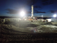 A white oil drill stands above several office trailers above an excavated pit about 50 meters wide and 5 meters deep. It is night. The scene is lit by 3 industrial light towers.
