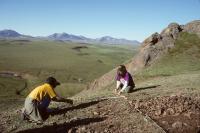Two people are working on a hillside with a helicopter flying in the background