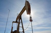 A low-angle shot captures the dark silhouette of a large oil pumpjack against a bright, sunlit sky. The sun is directly behind the pumpjack's horsehead, creating a brilliant flare and obscuring its details, while thin, wispy clouds streak across the blue sky. A thin antenna tower stands to the left of the machinery.