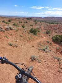 A point-of-view shot from a mountain bike looking down a winding red dirt trail in a vast, arid desert landscape
