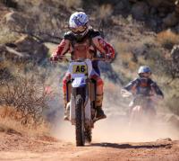 A rider sits on a motorcycle and wears a white helmet and red racing suite with dust kicking up behind in a desert landscape