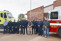 Firefighters in navy blue uniforms stand in front of two fire trucks; one green and one red fire truck