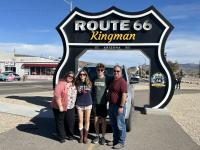 Four people stand in front of a large Route 66 Kingman roadside sign with a vintage vehicle display behind them on a sunny day.
