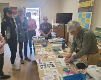 A woman demonstrates printmaking techniques to a small group gathered around a table filled with art prints and supplies in a bright room.