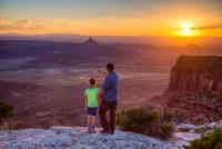 A father and daughter stand on an overlook with landscape features in the distance. 