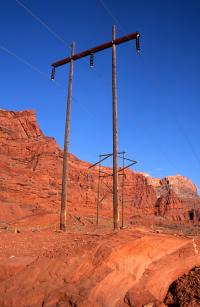 Wooden utility poles stand in a stark desert landscape against a deep blue sky. Behind the towers, massive, layered red rock formations and cliffs rise from the sandy, sun-drenched canyon floor.