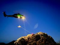 A helicopter hoists a litter from rescuers positioned on a rocky ridge during a nighttime rescue training exercise.