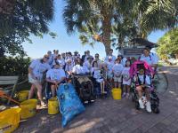 Group of people, including children and individuals in wheelchairs, gathered outdoors near palm trees with recycling buckets and bags.