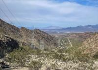 Power lines stretch across a rocky desert landscape with distant mountains under a partly cloudy sky.
