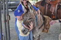 Child hugging a donkey wearing an American flag noseband, with a smiling adult behind and a saddled horse nearby.