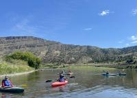 People in kayaks float on a river with hills in the background under a blue sky