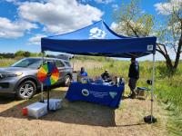 Outdoor event booth under a blue canopy with a colorful prize wheel and informational table near a silver SUV in a grassy field.