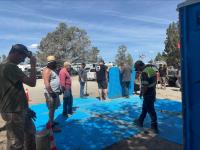 Group of people working outdoors on installing large blue interlocking panels on sandy ground near parked vehicles and trailers, with trees and flags visible in the background under a clear sky.